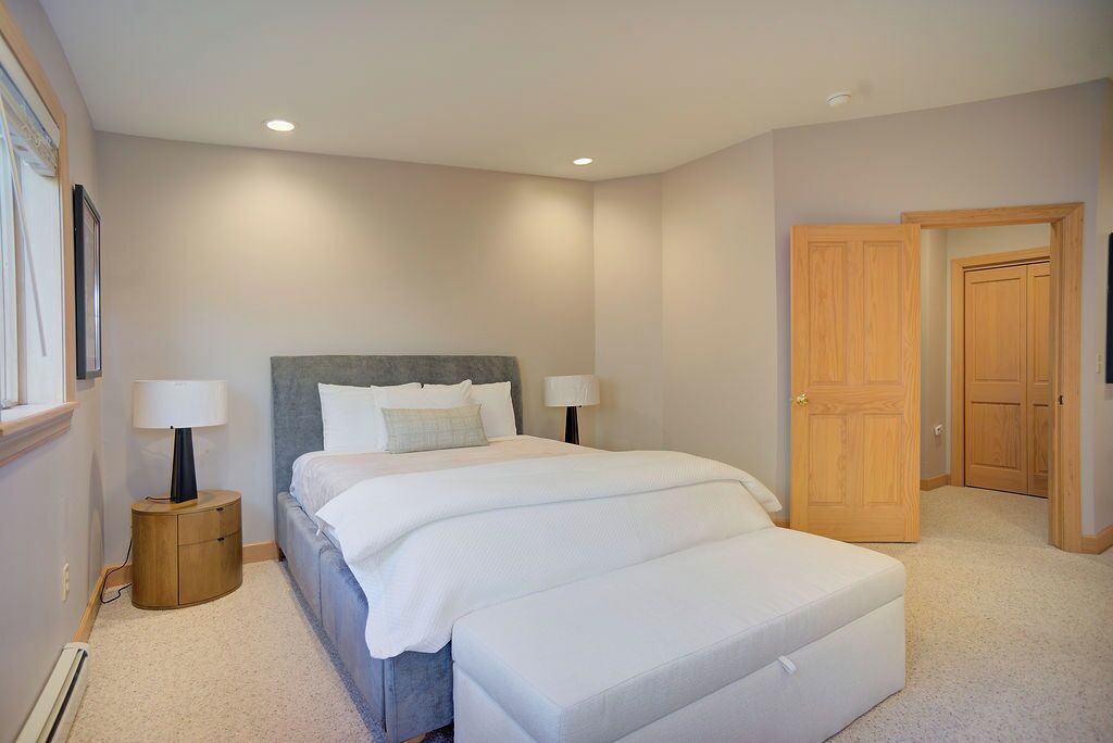 A bedroom with a large bed, two bedside tables with lamps, a wooden door, and a window with blinds.