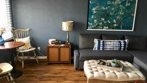 A living room with a grey sofa, a wooden coffee table with a plant, a blue and white patterned pillow, a lamp, and a framed floral painting on the wall.