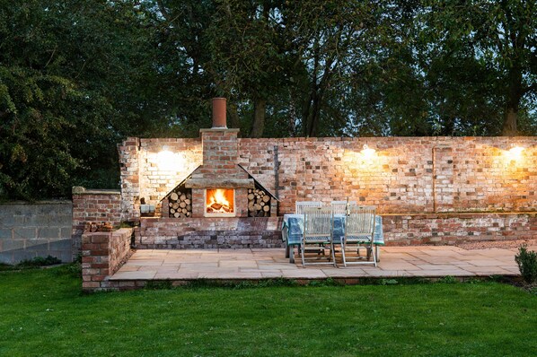 A brick outdoor fireplace with a fire, surrounded by a stone wall and a paved patio with two chairs. - The Hop Kiln and The Hayloft are two unique and attractive holiday lets (Monkland nr Leominster)