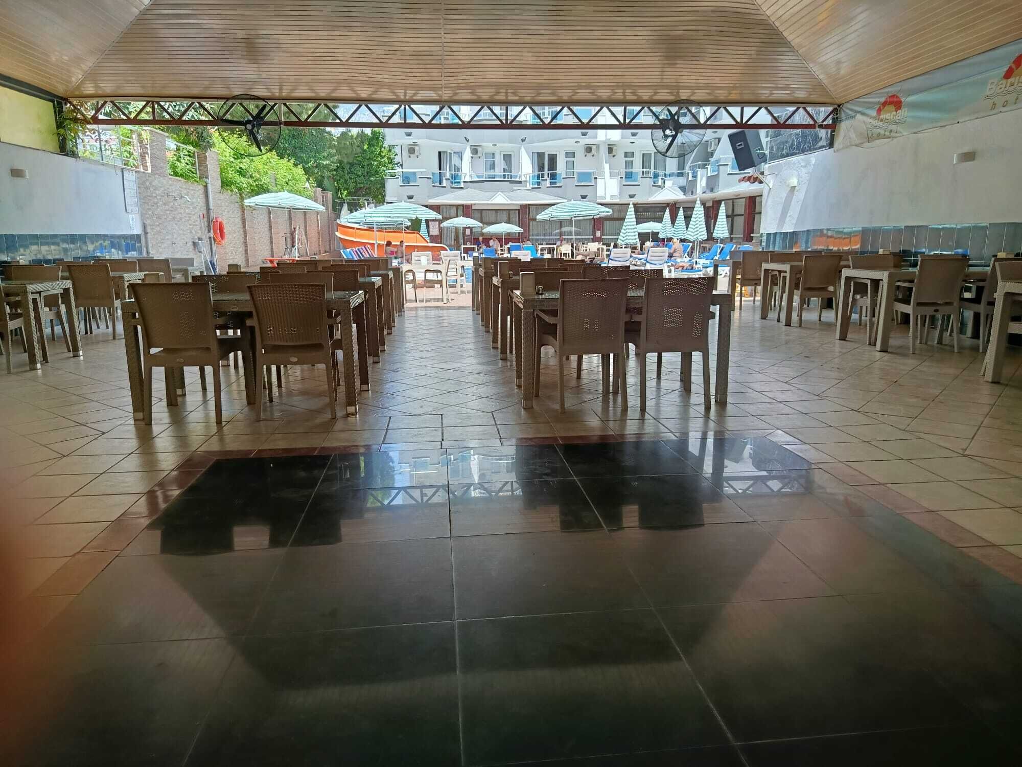 A spacious indoor dining area with rows of tables and chairs, a tiled floor, and a view of the sea and buildings in the background.