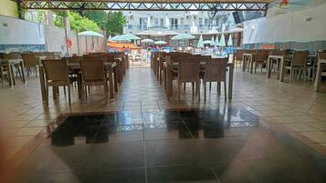 A spacious indoor dining area with rows of tables and chairs, a tiled floor, and a view of the sea and buildings in the background.