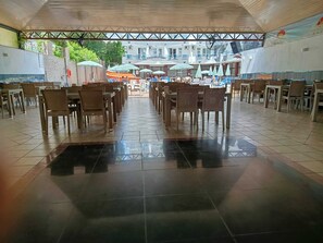 A spacious indoor dining area with rows of tables and chairs, a tiled floor, and a view of the sea and buildings in the background. - Bariscan Hotel (Alanya)