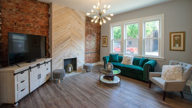A living room with a brick wall, a fireplace, a green sofa, a TV, and a modern chandelier. - Elegant & Cozy Escape - 5Beds/2.5Bath - Sleeps 10 (Pittsburgh)