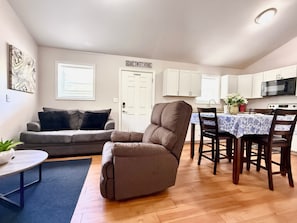 A living room with a gray sofa, a recliner, a dining table with chairs, a kitchen area with cabinets and appliances, and a door with a sign above it.