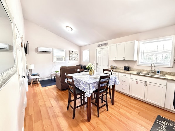 A compact kitchen with wooden flooring, white cabinets, a dining table with chairs, and a sofa.