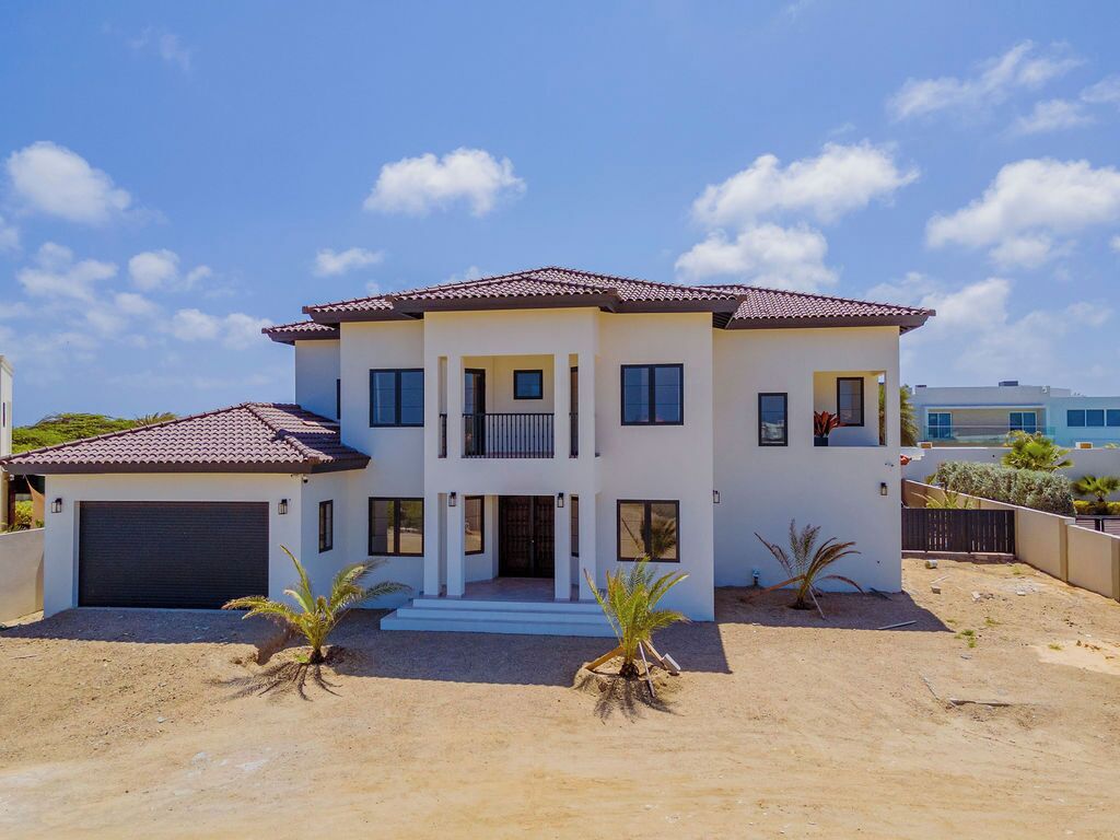 A two-story house with a balcony, a garage, and a sandy driveway.
