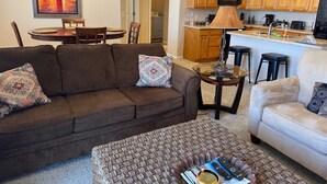 Ein Wohnzimmer mit einem braunen Sofa, einem weißen Sofa und einem gemusterten Hocker. Es befindet sich ein Esstisch mit Stühlen sowie eine Deckenleuchte. Die Küche ist sichtbar, mit Holzschränken und einer Mikrowelle.