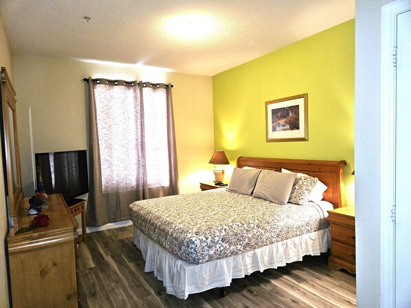 Un dormitorio con una cama de madera, un televisor sobre un mueble de madera, una mesita de noche con lámpara y una ventana con cortinas.