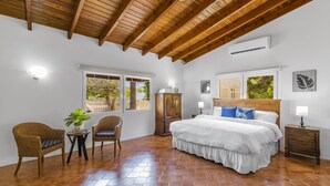 A bedroom with a wooden ceiling, a bed with white bedding, a nightstand, a chair, a small table with a plant, and a window with a view of trees.