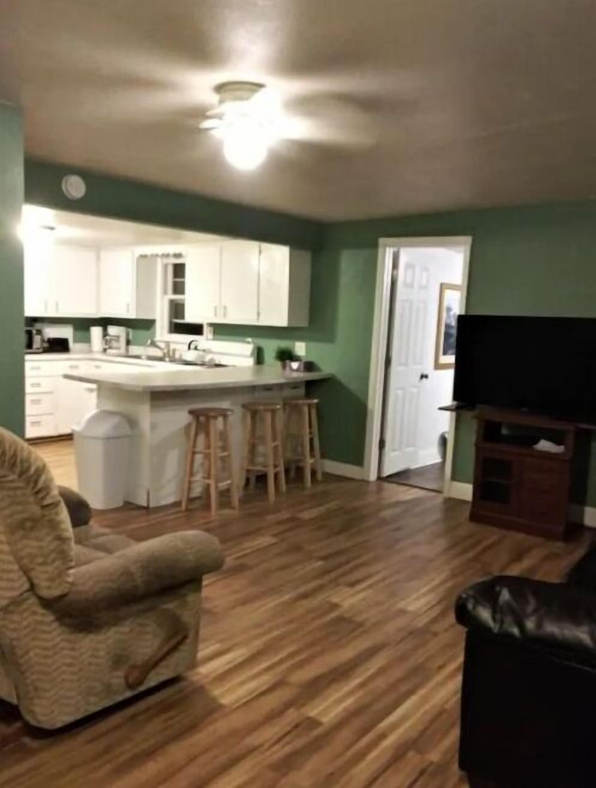 Un salon avec un plancher en bois, un canapé, un meuble pour la télévision et un coin cuisine doté de meubles blancs et d’un tabouret de bar.
