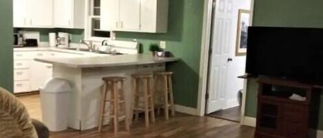Un salon avec un plancher en bois, un canapé, un meuble pour la télévision et un coin cuisine doté de meubles blancs et d’un tabouret de bar.