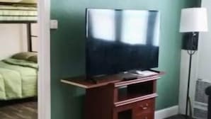 Una habitació amb terra de fusta, una televisió damunt d'un suport de fusta, un llit amb edredó verd i una làmpada.