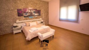 Quarto com cama, mesas de cabeceira, um banquinho, TV na parede e uma janela.