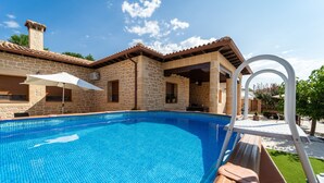 Piscina com paredes de pedra e teto telhado, rodeada por um alpendre com guarda-sol branco e um deck de madeira.