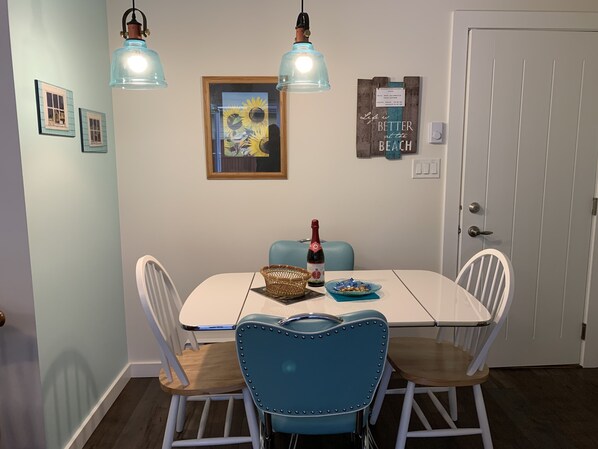 Un comedor con una mesa blanca, dos sillas azules, una botella de champán y un cuadro en la pared.