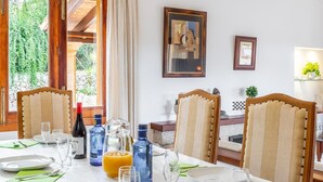 Comedor con una mesa dispuesta para cuatro, copas de vino, platos y una jarra de jugo de naranja. Hay dos sillas con tapicería a rayas. Un cuadro enmarcado está en la pared y una planta pequeña en un estante.