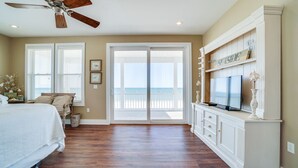 Une chambre avec un grand lit, un ventilateur de plafond, un téléviseur posé sur un meuble blanc et une vue sur la plage, visible à travers une porte-fenêtre coulissante.