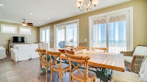 Un vaste espace de vie comprenant une table à manger en bois, des chaises en osier et une grande fenêtre donnant sur l’océan.