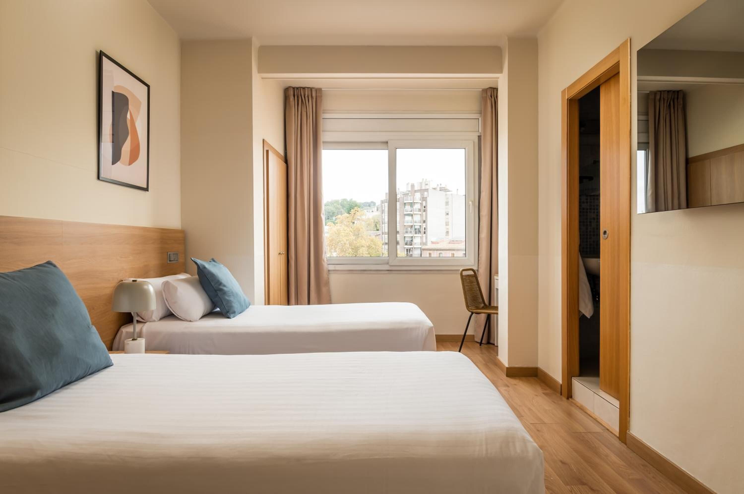 A hotel room with two beds, a window with curtains, a chair, and a wardrobe.
