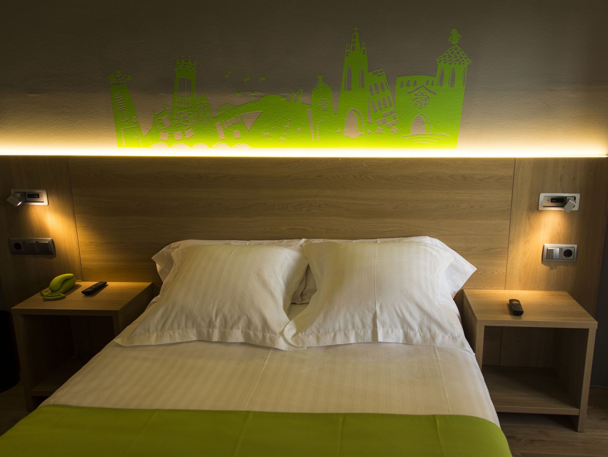 Habitación de hotel con cama, dos mesitas de noche, una lámpara de pared y una colcha verde.