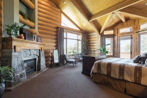 A log cabin bedroom with a stone fireplace, large windows, and a cozy bed.