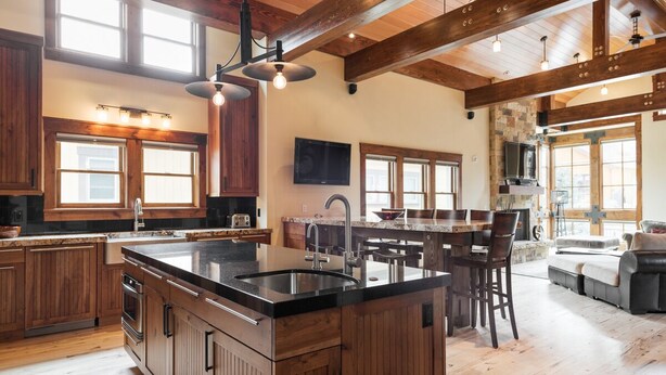 A spacious kitchen with wooden cabinets, a large island with a sink, and a dining area with a stone fireplace. - Woodside Villa (Park City)