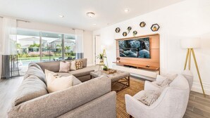 A modern living room with a sectional sofa, a flat-screen TV mounted on the wall, and a large window offering a view of the outdoors.