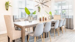 A modern dining room with a wooden table and grey upholstered chairs, surrounded by large windows and decorative plants.