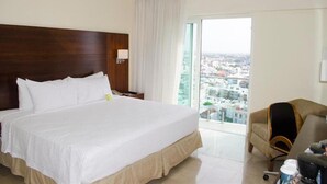 Une chambre d’hôtel avec un grand lit, une chaise, un bureau et une vue sur le paysage urbain, visible à travers une grande fenêtre.