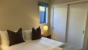 A neatly made bed with white and brown linens, a bedside lamp, and a window with a blue valance. - Central W ānaka Retreat | Close to the lake! (Wānaka)