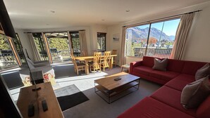 A spacious living area with a red sofa, wooden coffee table, and a dining table with chairs. Large windows offer a view of mountains and a balcony. - Central Wānaka Retreat | Close to the lake! (Wānaka)