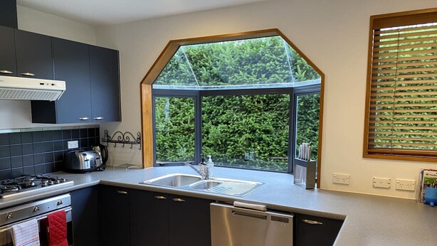 A modern kitchen with dark cabinets, a stainless steel dishwasher, a built-in oven, and a window with a view of greenery. - Central Wānaka Retreat | Close to the lake! (Wānaka)