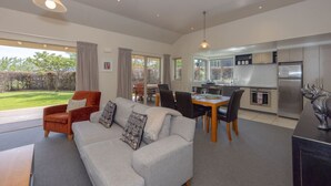 A modern living room with a sofa, dining table, and chairs, a kitchen with a refrigerator, and a view of a garden.