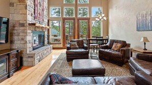 A spacious living room with a stone fireplace, leather sofas, a large window with a view of trees, and a dining area.