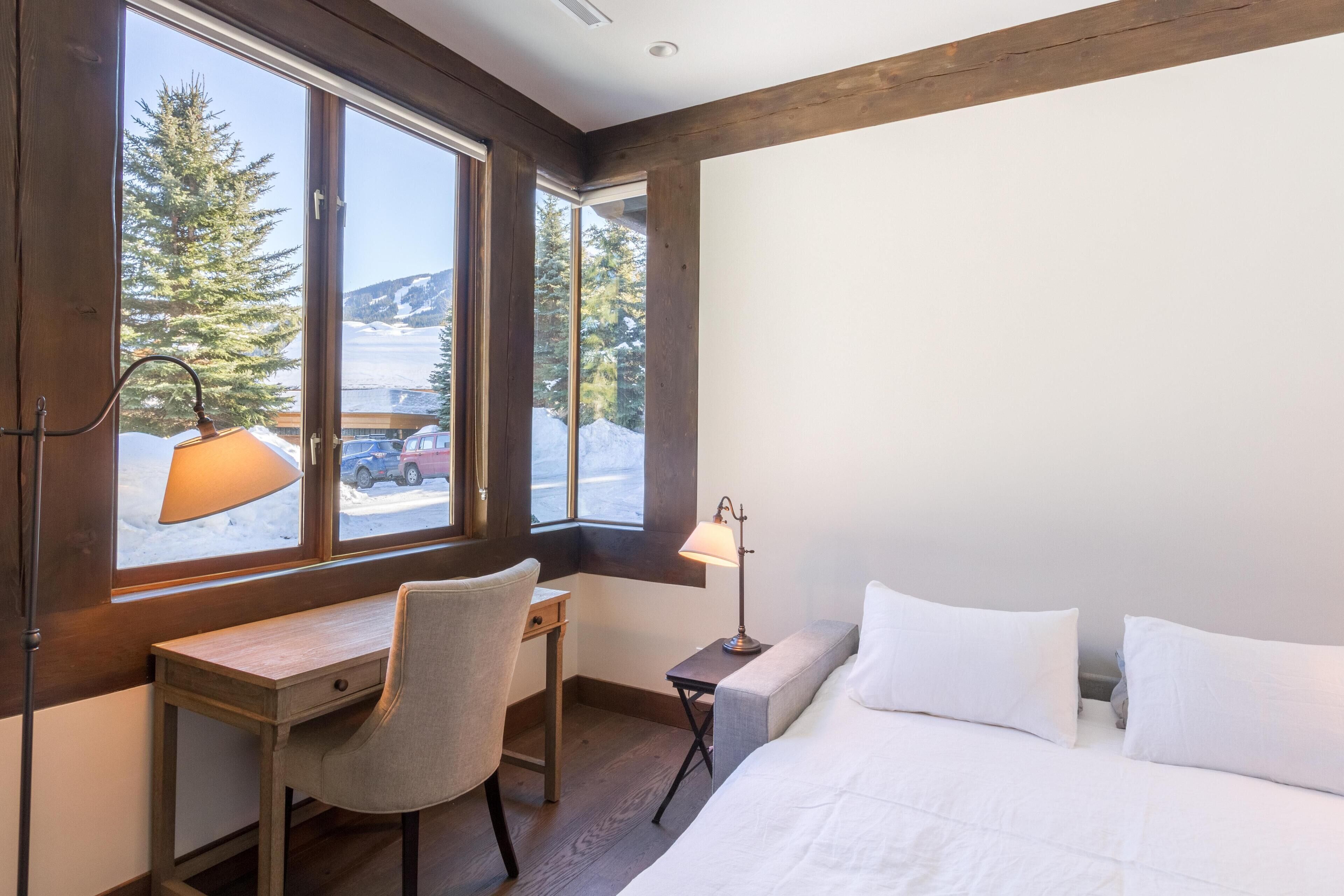 Une pièce avec un bureau en bois, une chaise, une table de chevet avec une lampe, un lit avec une literie blanche et une grande fenêtre donnant sur des arbres et des montagnes.