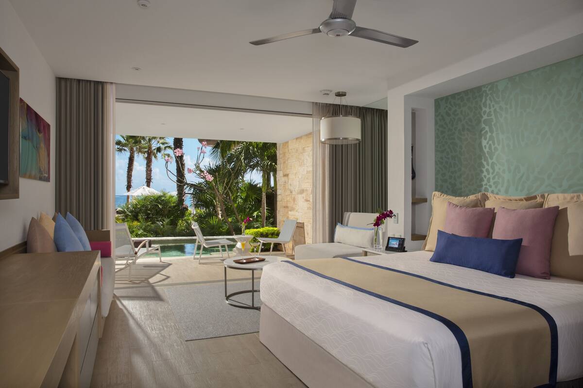 Une chambre d’hôtel avec un lit, un bureau, une chaise, une petite table et une vue sur une piscine et des palmiers.