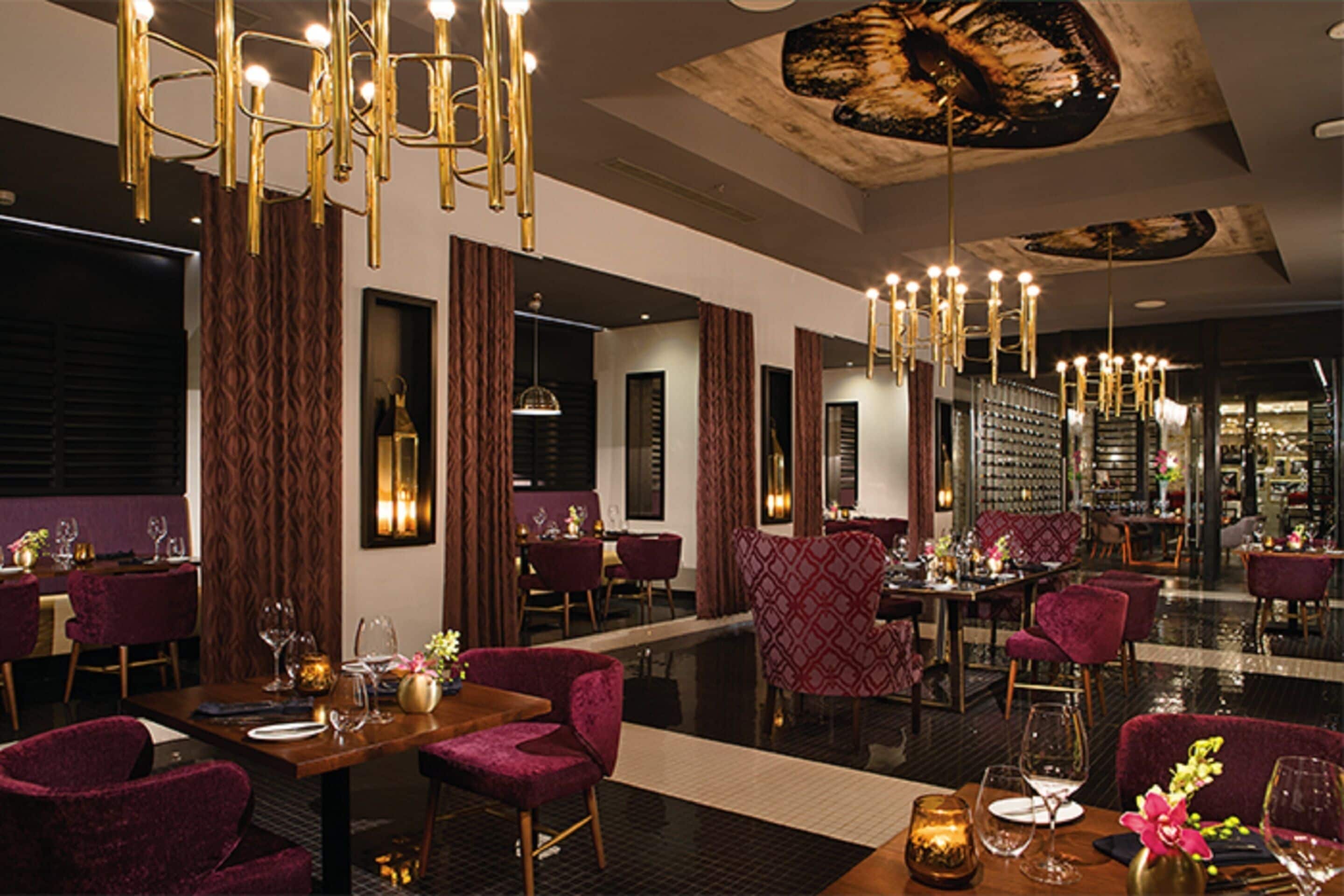 A modern restaurant with high ceilings, chandeliers, and red upholstered chairs.