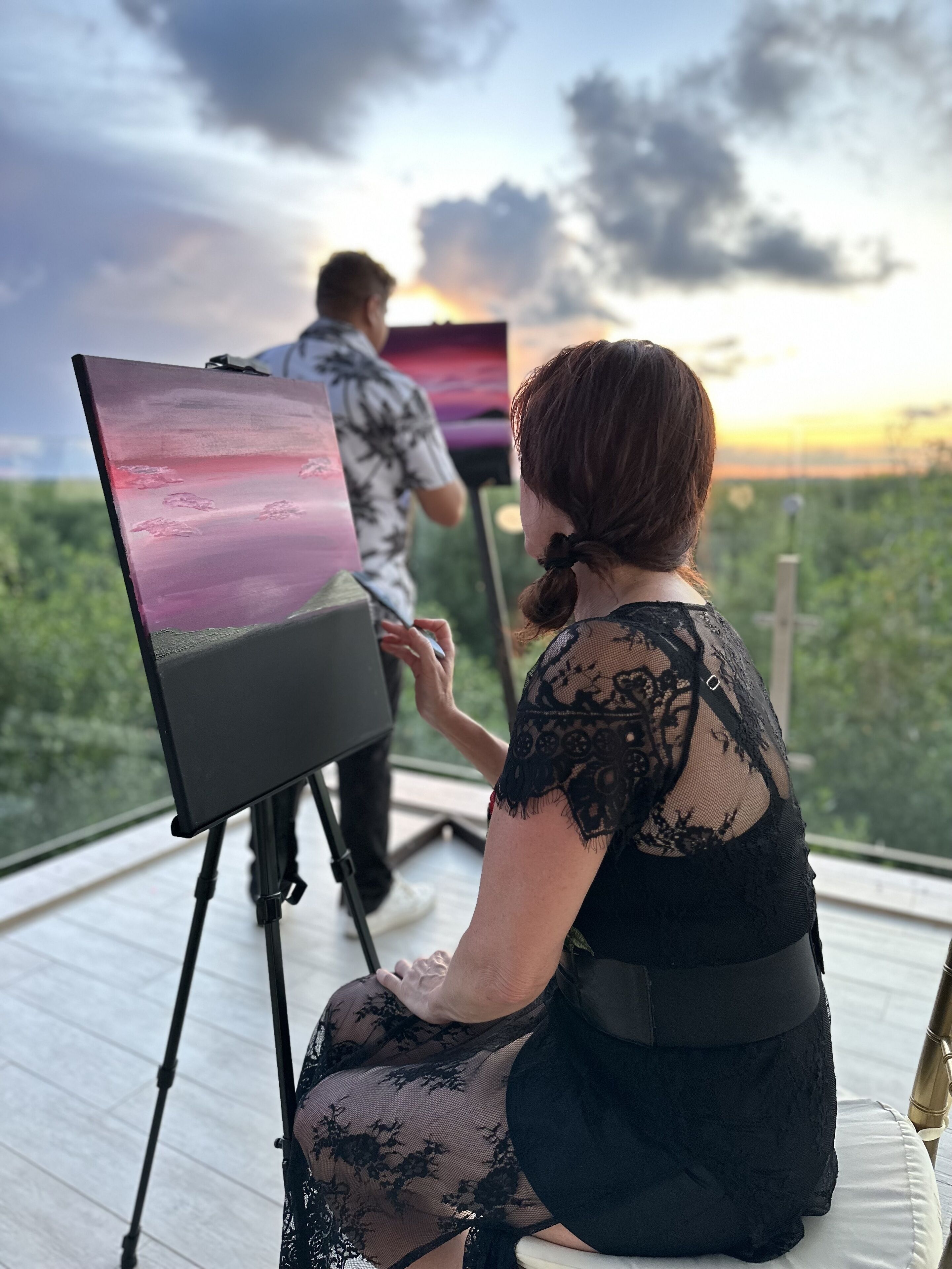 A woman sitting on a deck chair, facing a painting of a sunset. A man stands nearby, observing the painting.