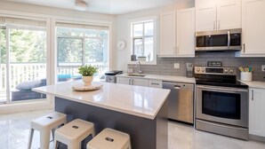 A modern kitchen with white cabinets, a large island with stools, stainless steel appliances, and a view of the outdoors. - Panoramic Views From This Gorgeous 2 Bed Property - Village Location (Whistler)