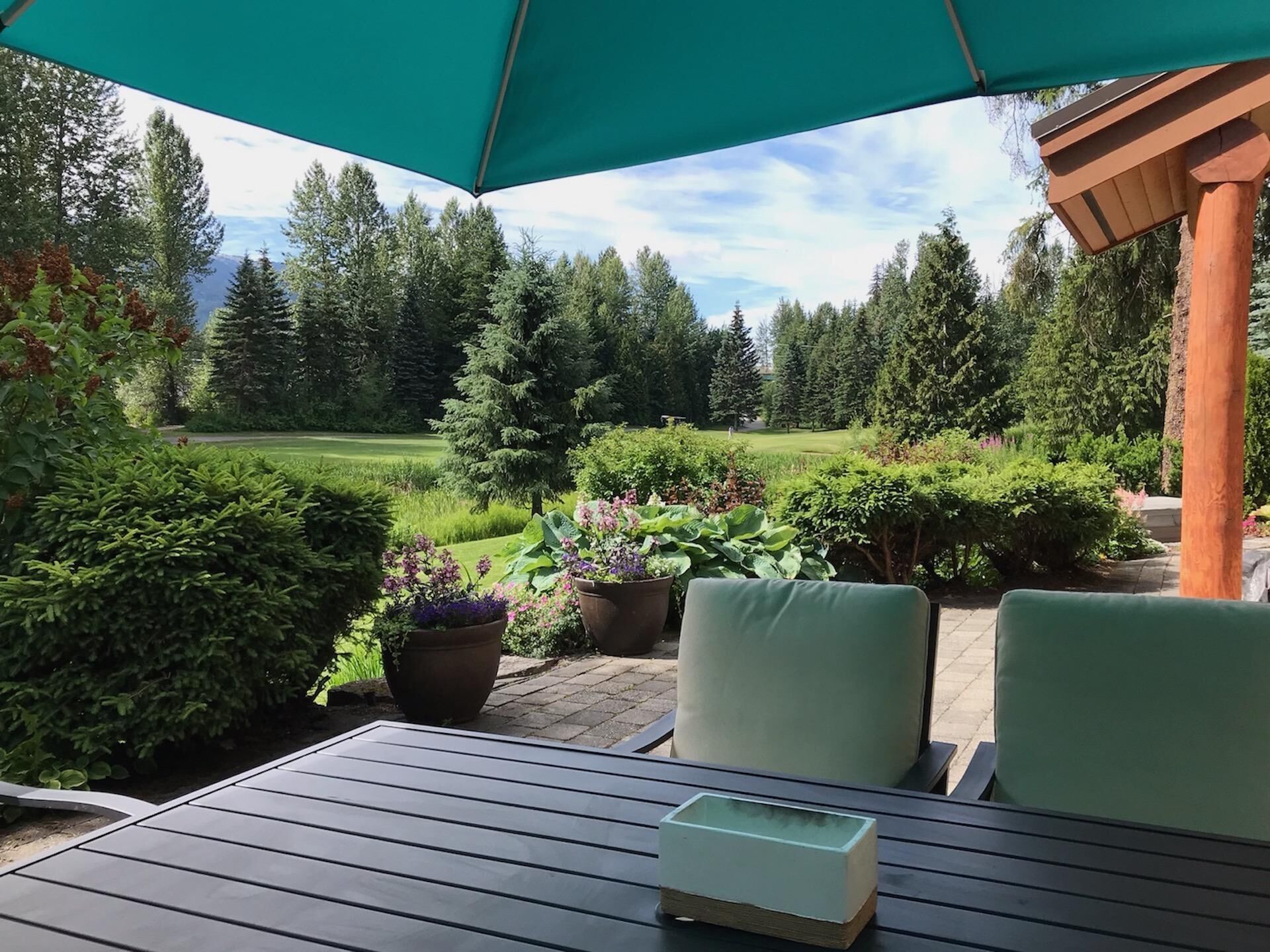 Un patio exterior con mesa y sillas, rodeado de plantas en macetas y vistas a un jardín exuberante y árboles.