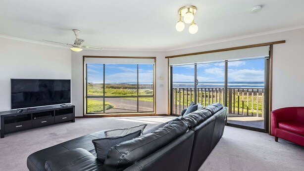 A modern living room with a large black leather sofa, a flat-screen TV, and a red chair. The room has large sliding glass doors leading to a balcony with a view of the ocean. - With South Beach views, a relaxed family home (PORT FAIRY)