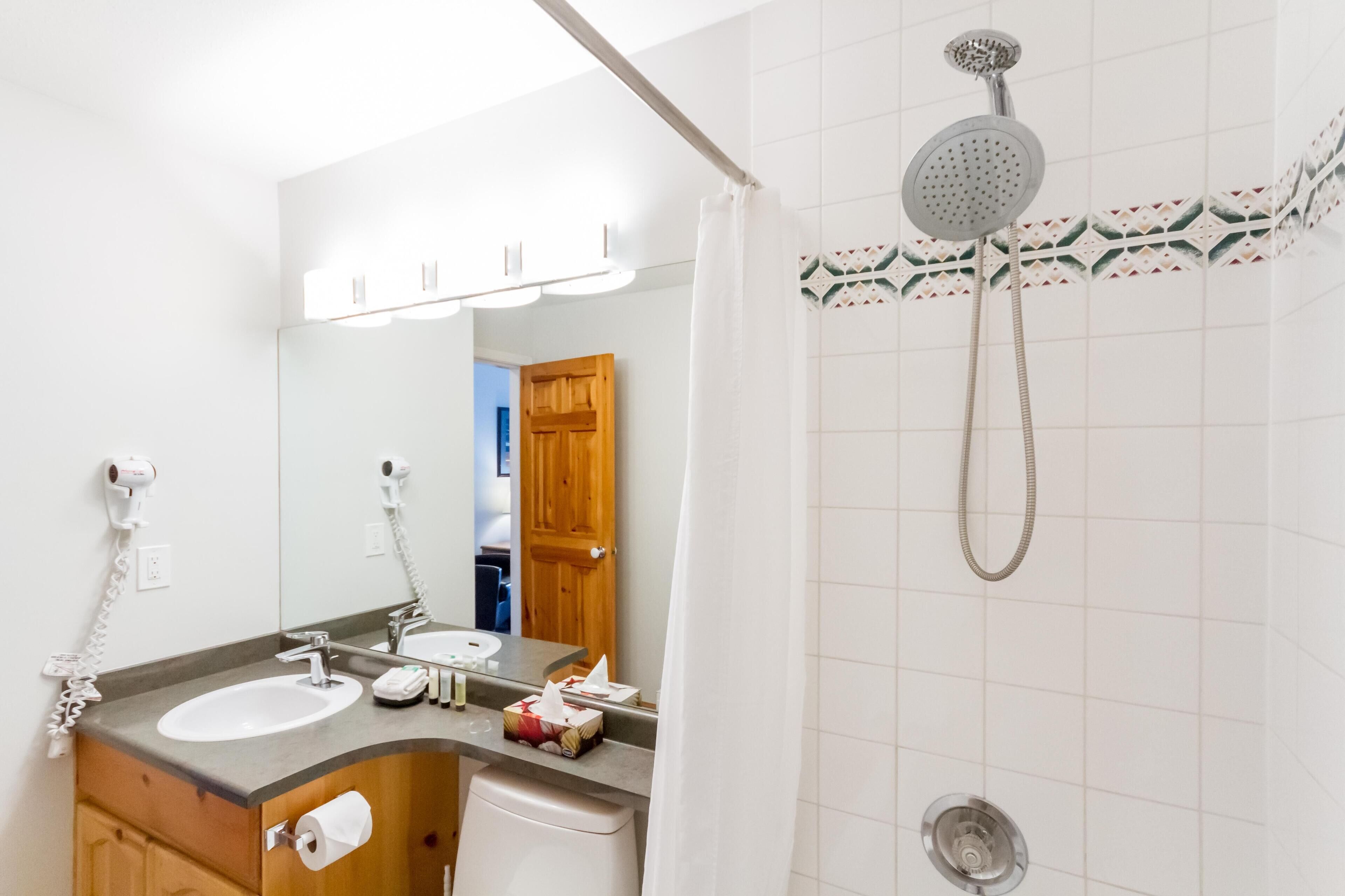 A bathroom with a sink, mirror, shower, and towel dispenser.