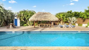 Une piscine entourée de chaises longues, un abri à toit de chaume et une végétation tropicale.