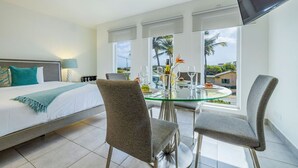 Une chambre d’hôtel avec un lit, une table à manger en verre entourée de chaises, un téléviseur fixé au mur et une vue sur les palmiers et les bâtiments à l’extérieur.