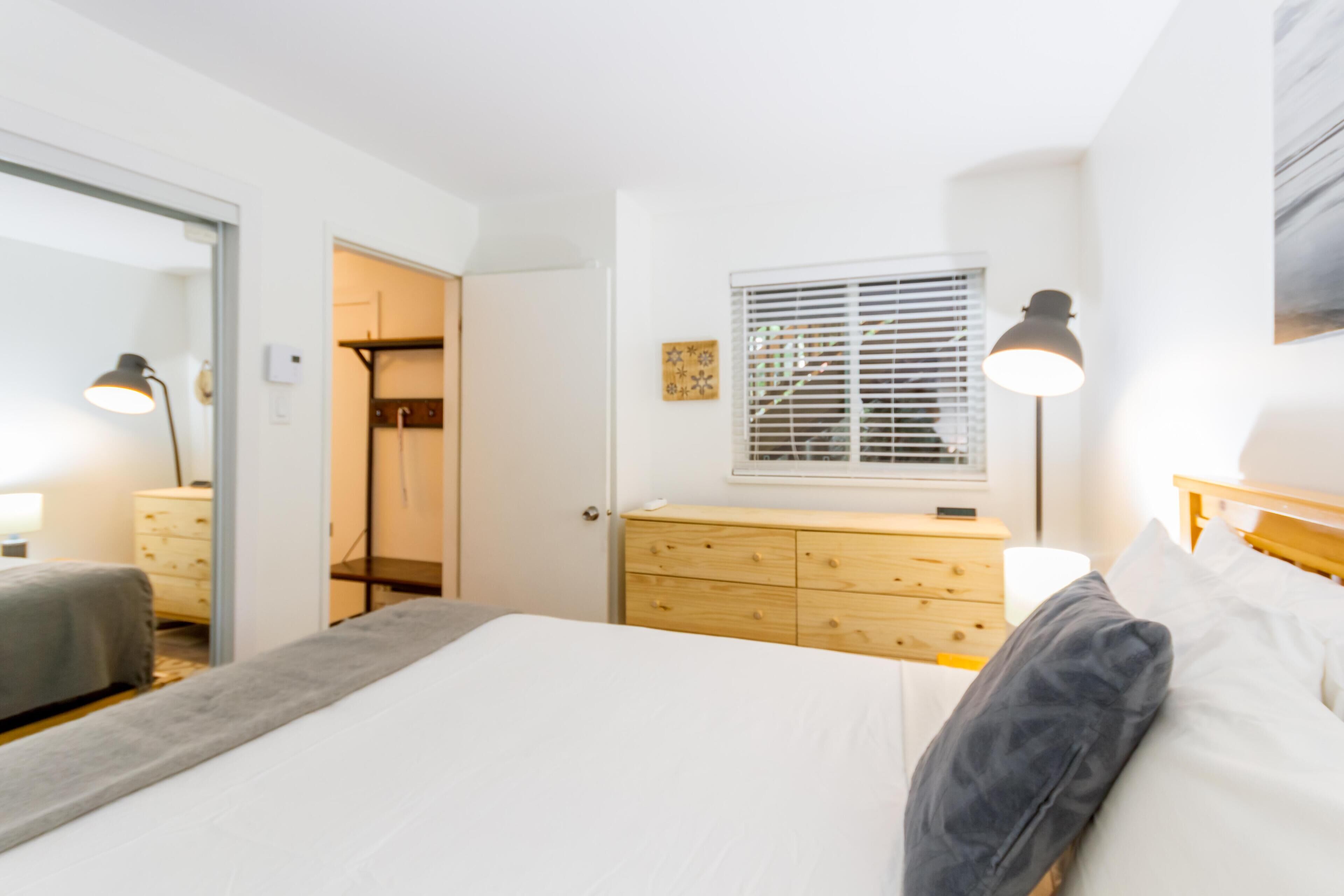 A bedroom with a bed, bedside table, lamp, dresser, and a window with blinds.
