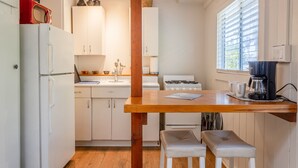 A compact kitchen with white cabinets, a wooden table, two stools, a stove, a coffee maker, and a window with blinds.