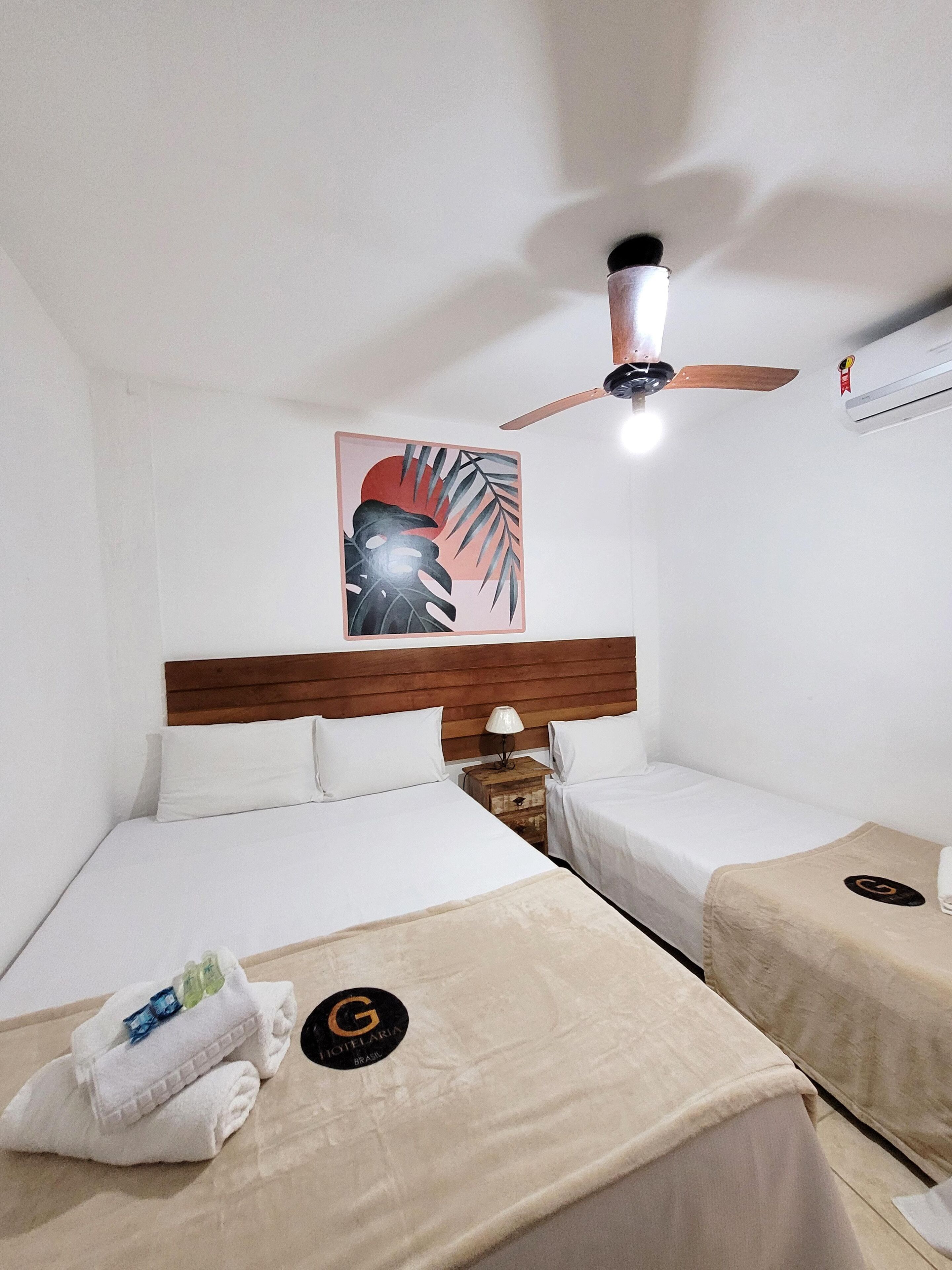 Een hotelkamer met twee bedden, een plafondventilator, een wandairconditioner en een schilderij met een tropisch thema.
