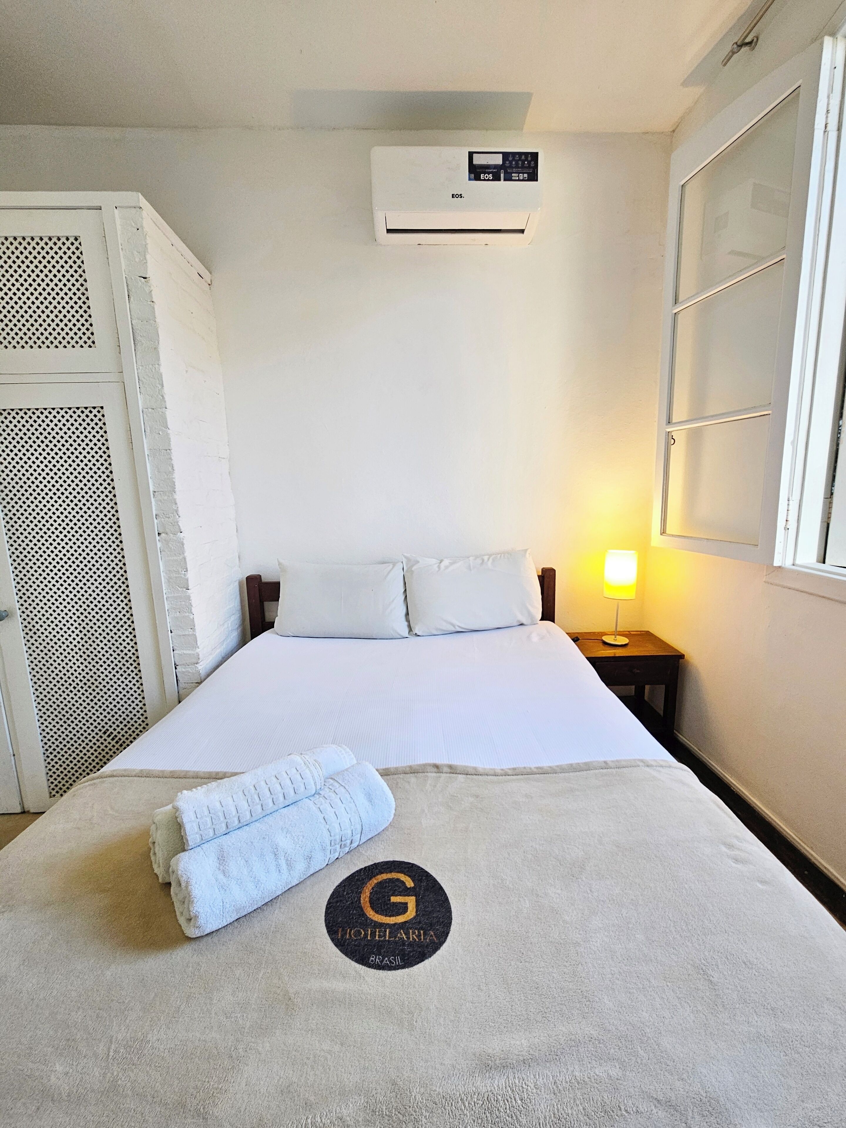 A hotel room with a bed, a nightstand with a lamp, an air conditioning unit, and a door with a mesh screen.