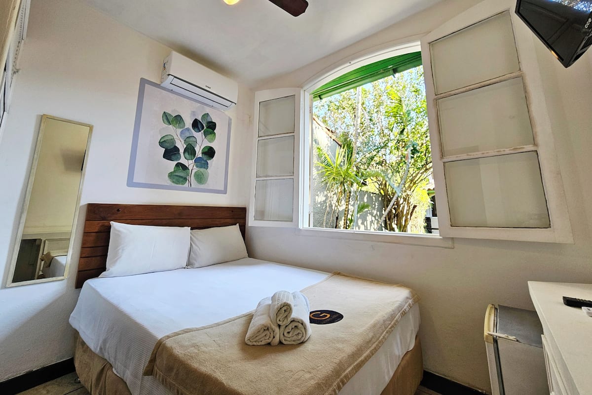 Ein Hotelzimmer mit Bett, Deckenventilator, Fenster mit Blick auf Bäume und einer Wand-Klimaanlage.