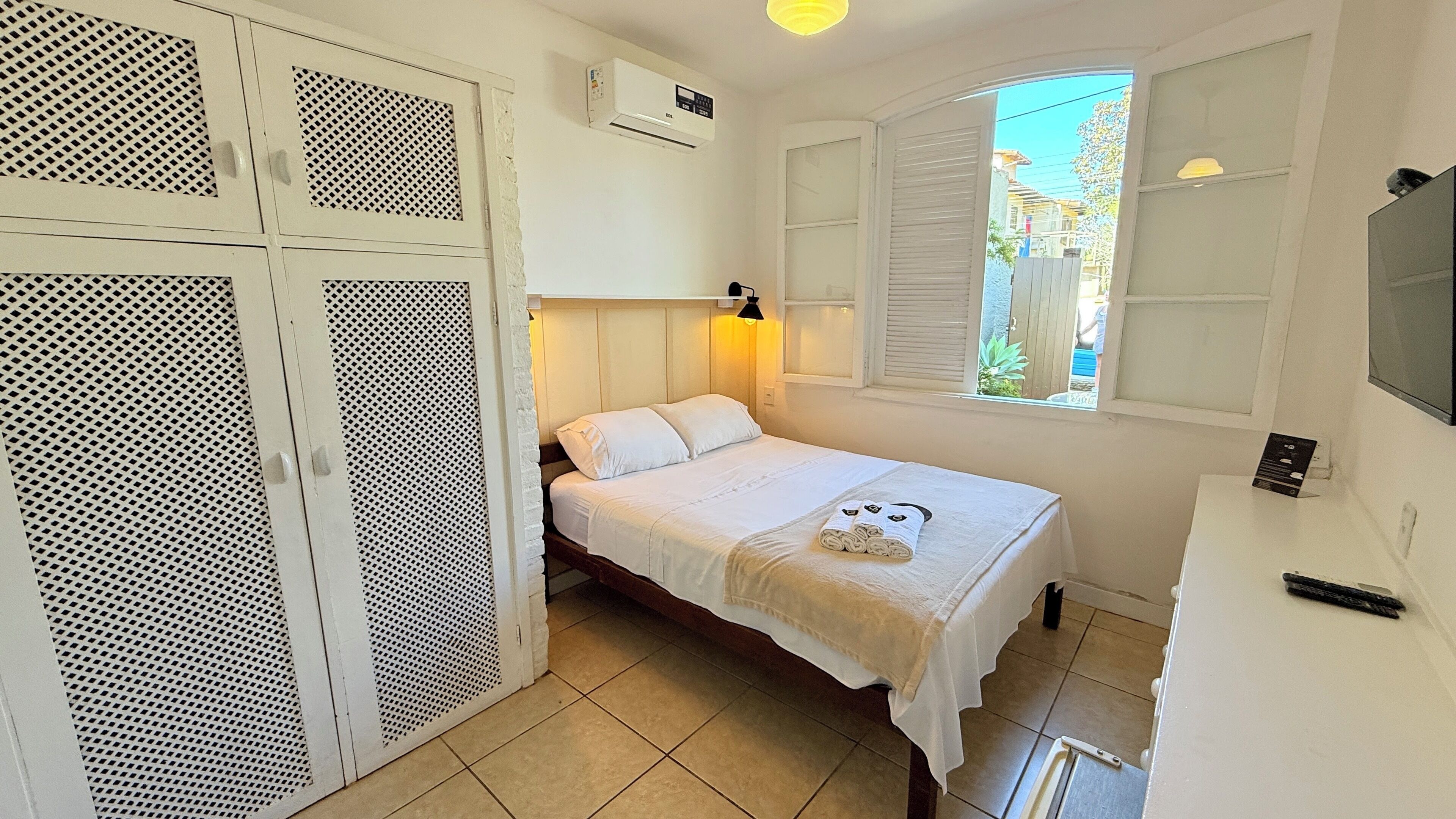 A small, well-lit bedroom with a bed, a window with shutters, a wardrobe, and an air conditioning unit.